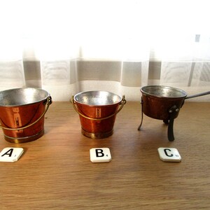 May include: Three miniature copper kitchenware items: a bucket with a handle, a smaller bucket, and a small pot with a handle and legs. The items are displayed on a wooden surface with white tiles labelled A, B, and C.
