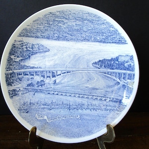 May include: A white ceramic plate with a blue and white illustration of a bridge over a river. The plate has a detailed drawing of the bridge, water, and surrounding landscape. The text "The Tranebergsbroar" is at the bottom.