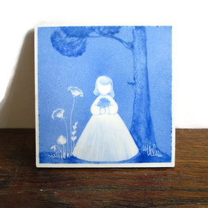 Op de afbeelding: Een blauwe en witte keramische tegel met een aquarel-illustratie van een meisje in een witte jurk dat een boeket bloemen vasthoudt. Het meisje staat voor een boom op een blauwe achtergrond. De tegel is gesigneerd met "Willems".