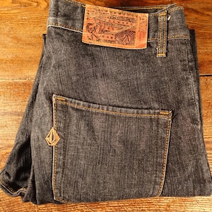 May include: A pair of dark blue denim jeans with a brown leather patch on the back pocket. The patch has a design and text on it.