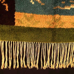 Op de afbeelding: Close-up van een geweven textiel met een gefranjerde rand. Het textiel heeft horizontale banden in teal, oranje en groen, met een donkerbruin gedeelte aan de linkerkant. De franje is lichtcr&egrave;me van kleur.