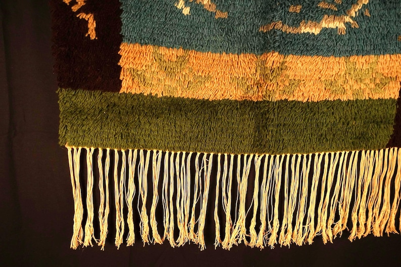 Op de afbeelding: Close-up van een geweven textiel met een gefranjerde rand. Het textiel heeft horizontale banden in teal, oranje en groen, met een donkerbruin gedeelte aan de linkerkant. De franje is lichtcr&egrave;me van kleur.