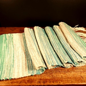 May include: Handwoven, rectangular table runner with a green and beige striped pattern. The runner is partially rolled up, revealing its texture and fringe edges. The runner is displayed on a wooden surface.