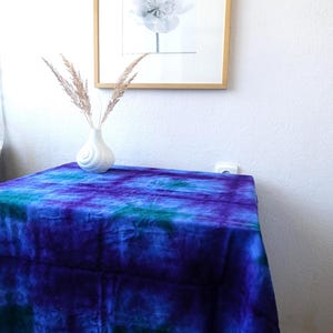 Może przedstawiać: Żywy obrus w stylu tie-dye w odcieniach niebieskiego i fioletowego pokrywa mały stół. Na wierzchu stoi biały wazon z suszonymi trawami. Oprawiony czarno-biały nadruk kwiatowy wisi na białej ścianie.