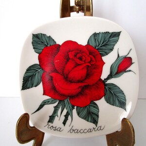 Op de afbeelding: Een vierkante, witte keramische plaat met een levendige rode roos en groene bladeren, met de tekst "rosa baccara". De plaat wordt gepresenteerd op een decoratieve messing standaard, die het bloemmotief benadrukt.