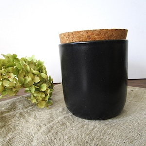 May include: A black ceramic jar with a cork lid. The jar is cylindrical and sits on a tan linen cloth. A sprig of dried green flowers is to the left of the jar.