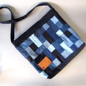 May include: A patchwork denim shoulder bag in various shades of blue. The bag features a rectangular shape with a long strap and a small, square, brown leather patch. The denim pieces are arranged in a geometric pattern.