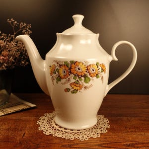 Könnte beinhalten: Weiße Keramik-Teekanne mit einem Blumendesign mit gelben und orangefarbenen Blumen. Die Teekanne hat einen Henkel und einen Ausguss. Sie steht auf einer weißen Häkelunterlage.