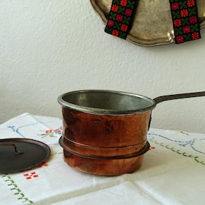 Puede incluir: Una cacerola de cobre con un mango largo y una tapa a juego descansa sobre un mantel floral. La olla tiene un mango y un borde de metal oscuro. La tapa se coloca a la izquierda de la olla. Una bandeja decorativa cuelga en el fondo.