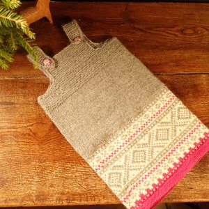 Puede incluir: Un calcetín navideño de punto gris con un patrón geométrico en blanco y rosa. El calcetín tiene dos correas con botones florales rosas. El borde inferior es de color rosa brillante. El calcetín está sobre una superficie de madera.