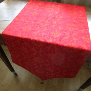 May include: A red table runner with a floral pattern. The runner is made of a textured fabric and is draped over a wooden table. The runner's color is a vibrant red with a subtle orange undertone, adding warmth to the dining area.