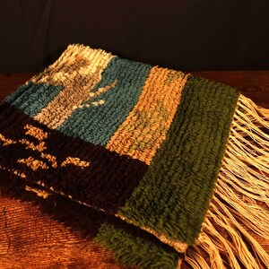 Op de afbeelding: Een gevouwen, geweven textiel met een getextureerd oppervlak en gefranjerde randen. Het textiel heeft een colorblock-ontwerp met secties in teal, bruin, groen en zwart met een lichtgekleurd patroon. Het textiel is waarschijnlijk een decoratieve sprei of deken.
