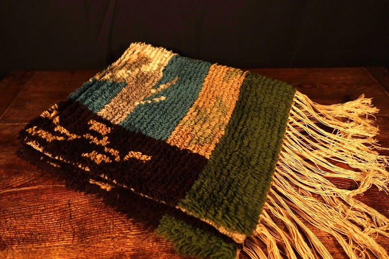 Op de afbeelding: Een gevouwen, geweven textiel met een getextureerd oppervlak en gefranjerde randen. Het textiel heeft een colorblock-ontwerp met secties in teal, bruin, groen en zwart met een lichtgekleurd patroon. Het textiel is waarschijnlijk een decoratieve sprei of deken.