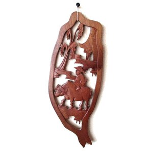 Op de afbeelding: Een bruine houten wanddecoratie met een gegraveerd tafereel van een persoon die op een waterbuffel door een bos rijdt.