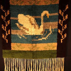 Op de afbeelding: Een geweven wandkleed met een zwaan op een blauwgroene achtergrond, met bruine, bruingele en groene horizontale strepen. De zwaan is cr&egrave;mekleurig met een oranje snavel. De onderkant heeft een franje.