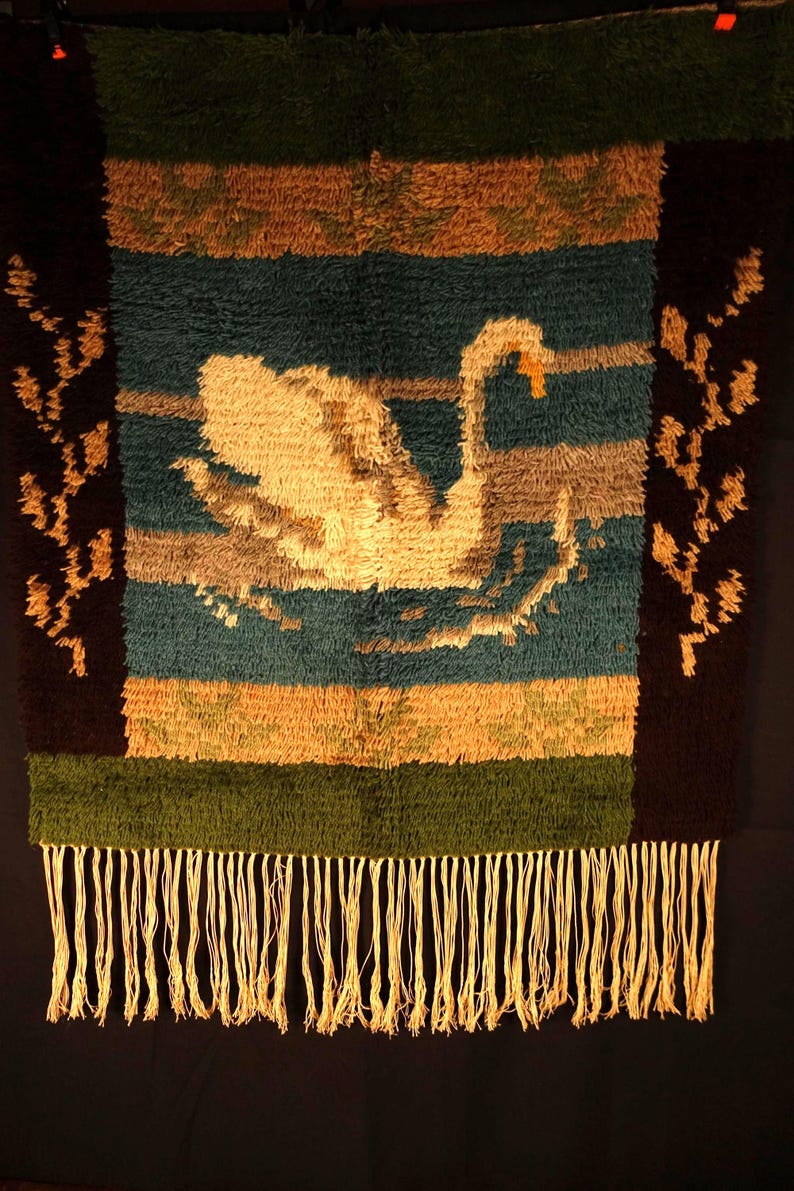Op de afbeelding: Een geweven wandkleed met een zwaan op een blauwgroene achtergrond, met bruine, bruingele en groene horizontale strepen. De zwaan is cr&egrave;mekleurig met een oranje snavel. De onderkant heeft een franje.