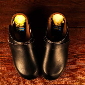 May include: A pair of black leather clogs with wooden soles, set against a warm, wooden background. The clogs have a classic design with a strap across the top. The interior shows a yellow label with text and a blue and yellow emblem.