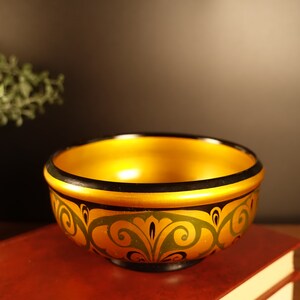 Pode incluir: Uma tigela decorativa de madeira com interior dourado e borda preta. O exterior apresenta uma base dourada com um desenho floral preto e verde. A tigela está em cima de um livro vermelho.