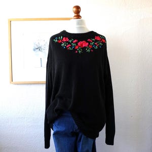 May include: Black sweater with a crew neck and long sleeves. The sweater has an embroidered floral design across the neckline, with red roses, green leaves, and small blue flowers. The sweater is displayed on a mannequin with blue jeans.