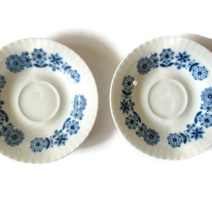 May include: Two white ceramic saucers with a blue floral pattern around the rim. The floral design features various stylized flower shapes. The saucers have a slightly ridged edge and a central indentation for a cup.