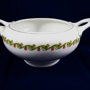 May include: White ceramic serving bowl with two handles and a decorative border. The border features a green and red floral pattern. The bowl has a gold rim and sits on a small pedestal base. The bowl is likely vintage.