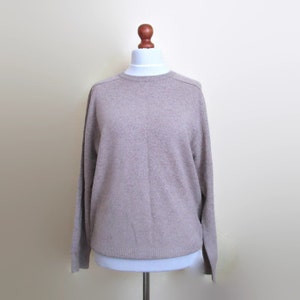 May include: A pale brown crew neck sweater with long sleeves. The sweater is displayed on a white mannequin with a wooden head. The sweater has a simple, classic design.