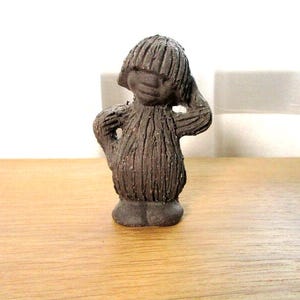 May include: A dark brown, textured ceramic figurine of a small, standing creature with a rounded body, short arms, and a bowl-cut hairstyle. The surface of the figure features vertical, etched lines throughout, giving it a fibrous appearance.