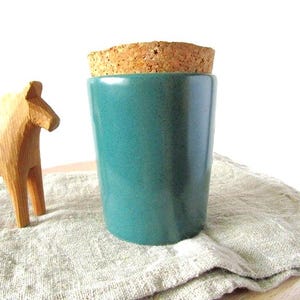 May include: A teal ceramic tumbler with a cork lid sits on a linen cloth. A carved wooden horse figurine is to the left. The tumbler is cylindrical and has a smooth, glossy finish.