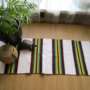 May include: A rectangular table runner featuring a repeating striped pattern in white, dark brown, bright yellow, and teal. The fabric has a textured, woven appearance with fringe detailing at both ends, laid out horizontally on a flat surface.