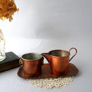 May include: A set of copper creamer and sugar bowl with a matching tray. The creamer and sugar bowl have a hammered finish and are on a lace doily.