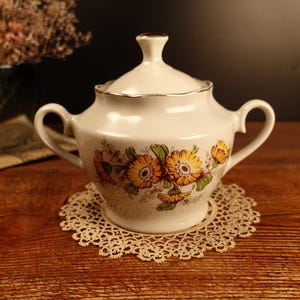 Könnte beinhalten: Weiße Porzellan-Zuckerdose mit Deckel und zwei Henkeln. Die Dose ist mit einem floralen Muster aus gelben und orangefarbenen Blumen auf weißem Hintergrund verziert.