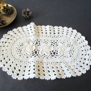 Op de afbeelding: Crèmekleurige, ovale gehaakte doily. Het midden heeft een bloemenpatroon, omgeven door een openwerkpatroon en een geschulpte rand. De doily is gemaakt van een zacht, getextureerd garen, perfect om een vintage touch aan de woondecoratie toe te voegen.