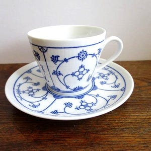 May include: A white porcelain teacup and matching saucer featuring a delicate blue floral and geometric pattern. The cup has a curved handle and a flared rim, while the saucer displays a circular arrangement of the same blue botanical motifs.