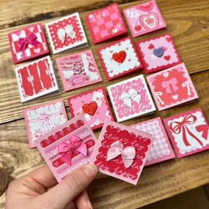 Könnte beinhalten: Eine Sammlung von Valentinstagskarten in verschiedenen Rosa- und Rottönen, mit Herzen, Schleifen und dem Text "Valentine's Day" und "love". Einige Karten haben strukturierte Elemente. Die Karten werden auf einer Holzoberfläche präsentiert.