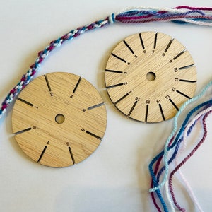 Op de afbeelding: Twee ronde houten gereedschappen met genummerde sleuven voor het maken van vriendschapsarmbanden. De gereedschappen zijn gemaakt van lichtbruin hout en hebben een natuurlijke nerf. Een meerkleurig vriendschapsarmband is gedeeltelijk gemaakt en ligt op een wit oppervlak.