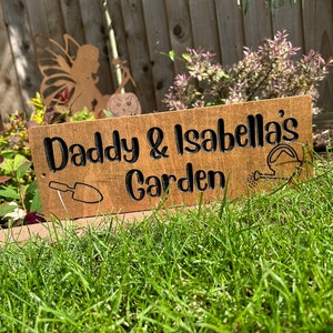 Könnte beinhalten: Ein Holzschild mit dem Text "Daddy & Isabella's Garden" in schwarzen Buchstaben. Das Schild ist mit einer kleinen Schaufel und einer Gießkanne verziert.