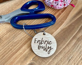 May include: A wooden tag with the text "Fabric only" hangs from a silver chain. Blue-handled scissors and a pink floral pin cushion with red pins are also visible. The items are on a wooden surface.