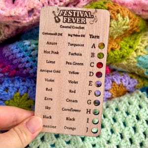 Könnte beinhalten: Eine Holz-Farbkarte für Coastal Crochet mit der Aufschrift "Festival Fever". Sie listet Garnfarben wie Azure, Hot Pink und Lime mit entsprechenden Farbmustern auf. Die Karte wird über einer bunten Häkeldecke gehalten.