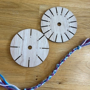 Op de afbeelding: Twee ronde houten gereedschappen met de nummers 1 tot en met 16 erop gemarkeerd. De gereedschappen worden gebruikt om vriendschapsarmbanden te maken. Een veelkleurige vriendschapsarmband ligt op een houten oppervlak.