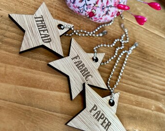 May include: Three wooden star-shaped tags with the words "THREAD", "FABRIC", and "PAPER" engraved on them. Each tag has a silver chain attached.