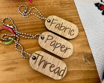 May include: Three wooden tags with metal chains, labeled "Fabric", "Paper", and "Thread". The tags are on a wooden surface with a pincushion and a pair of scissors in the background.