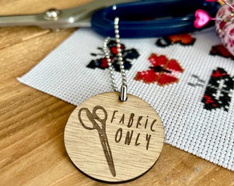 May include: A round wooden tag with a silver chain attached. The tag has a laser-engraved design of a pair of scissors and the text "FABRIC ONLY".