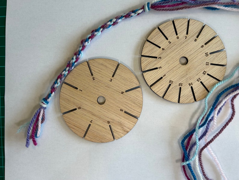 Op de afbeelding: Twee ronde houten gereedschappen met genummerde sleuven voor het maken van vriendschapsarmbanden. De gereedschappen zijn gemaakt van lichtbruin hout en hebben een centraal gat. De nummers op de gereedschappen vari&euml;ren van 1 tot 16 en van 1 tot 12. Er zijn ook strengen roze, blauwe en witte garen.