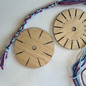 Op de afbeelding: Twee ronde houten gereedschappen met genummerde sleuven voor het maken van vriendschapsarmbanden. De gereedschappen zijn gemaakt van lichtbruin hout en hebben een centraal gat. De nummers op de gereedschappen vari&euml;ren van 1 tot 16 en van 1 tot 12. Er zijn ook strengen roze, blauwe en witte garen.