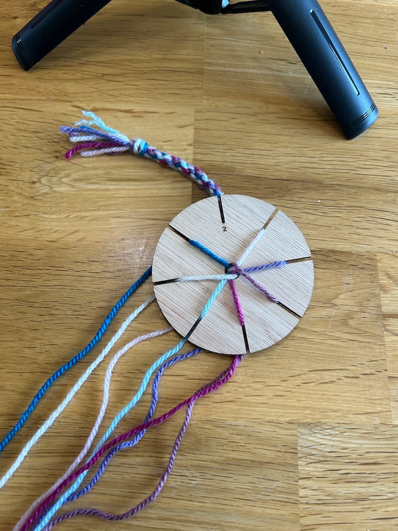 Op de afbeelding: Een houten weefgetouw met acht gleuven voor garen, gebruikt voor het maken van vriendschapsarmbanden. Het weefgetouw heeft zes draden garen bevestigd, in de kleuren roze, wit, lichtblauw, donkerblauw, paars en turquoise.