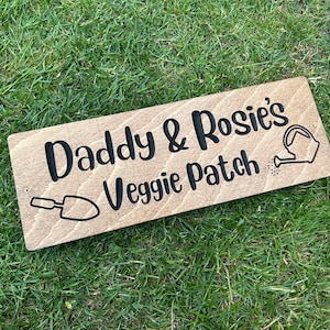 May include: A rectangular wooden sign with the words "Daddy & Rosie's Veggie Patch" in black. It features a trowel and watering can graphic. The sign is made of light wood and is set on green grass.