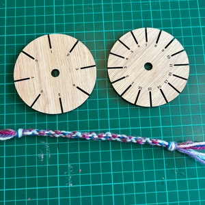 Op de afbeelding: Twee ronde houten gereedschappen met genummerde markeringen voor het maken van vriendschapsarmbanden. Een veelkleurig vriendschapsarmband ligt op een groene snijmat.