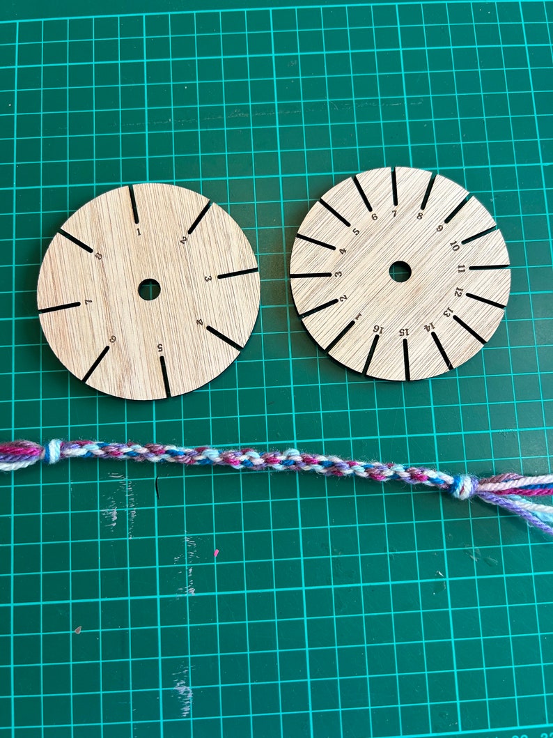 Op de afbeelding: Twee ronde houten gereedschappen met genummerde markeringen voor het maken van vriendschapsarmbanden. Een veelkleurig vriendschapsarmband ligt op een groene snijmat.