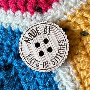 Pode incluir: Um botão de madeira com o texto "Made by Kat's-in-Stitches" e três botões pretos no centro.