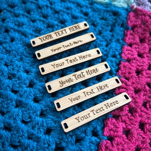 Könnte beinhalten: Sechs rechteckige Holzanhänger mit den eingravierten Texten "YOUR TEXT HERE" und "Your Text Here". Die Anhänger haben kleine Löcher an jedem Ende und werden auf einer farbenfrohen Häkeldecke in Blau-, Türkis- und Rosatönen präsentiert.